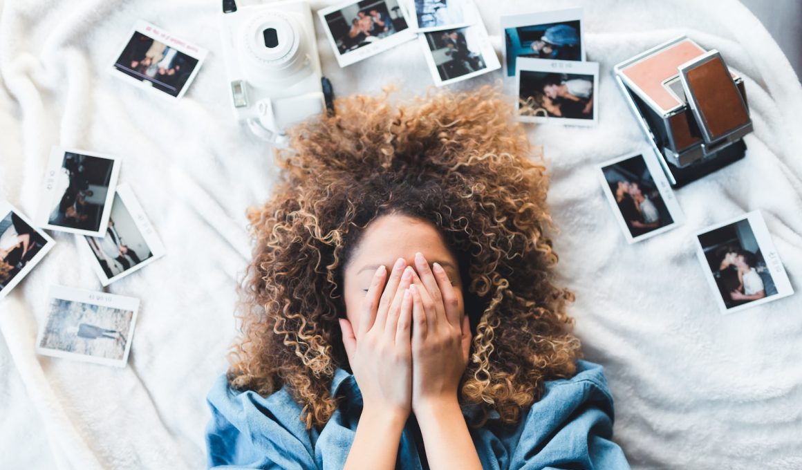 woman lying on bed covering her face surrounded by photos and white camera