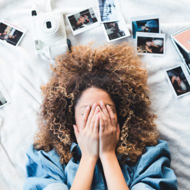 woman lying on bed covering her face surrounded by photos and white camera