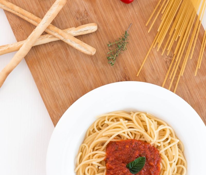 white ceramic bowl with pasta and red tomato on brown wooden table