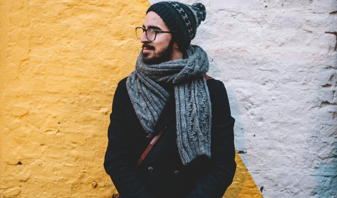 man in black coat with grey scarf standing on front of white and yellow painted wall