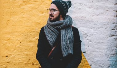 man in black coat with grey scarf standing on front of white and yellow painted wall