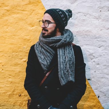 man in black coat with grey scarf standing on front of white and yellow painted wall