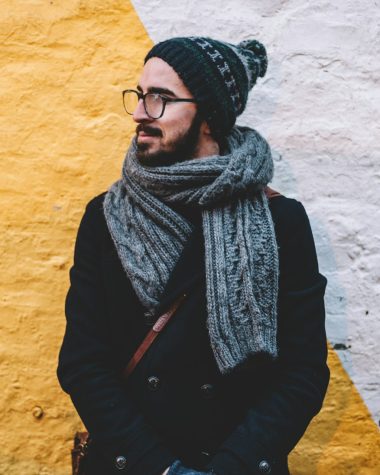 man in black coat with grey scarf standing on front of white and yellow painted wall